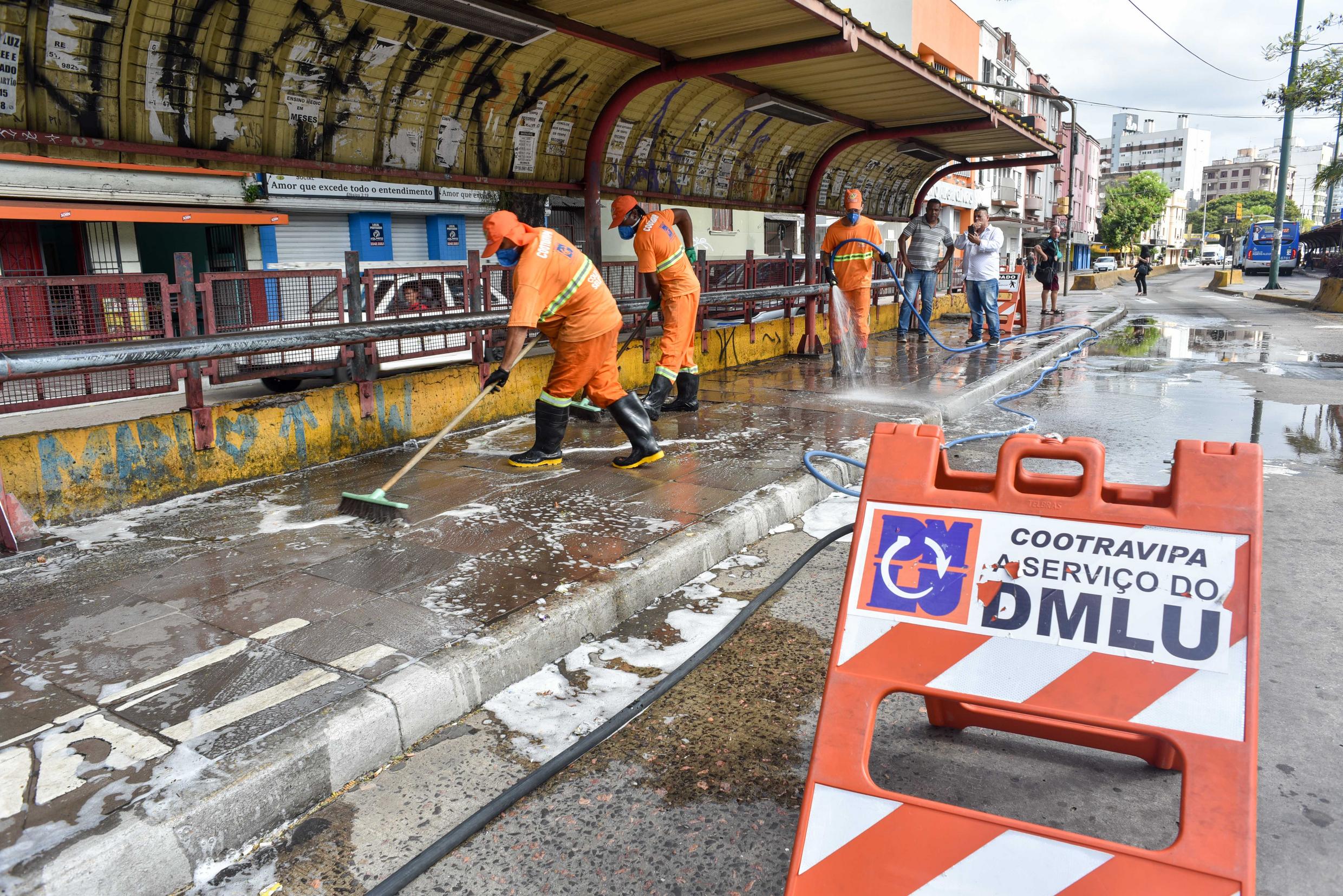 A imagem mostra garis realizando operação de lavagem e desinfecção em uma parada de ônibus em Porto Alegre.