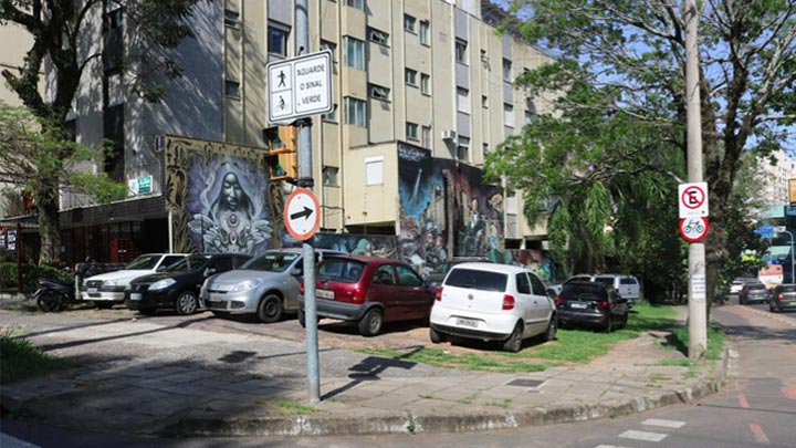 Terreno abandonado, com carros estacionados sobre o calçamento e a grama. Na frente do terreno há um semáforo, e ao lado um poste com placas de trânsito.