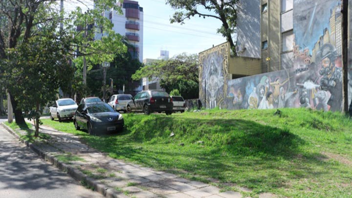 Terreno abandonado, com carros estacionados sobre a grama. Nos fundos do terreno, um prédio antigo com um graffiti desbotado na parede
