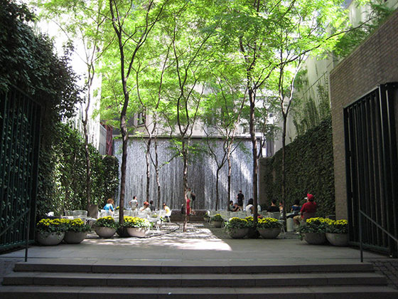 Pocket park entre prédios, composto por algumas árvores, vasos de plantas, mesas e cadeiras para os transeuntes e uma pequena cachoeira artificial ao fundo