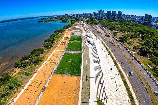 Imagem aérea de Porto Alegre com o Guaíba à esquerda em tons de azul e marrom, parte 3 da orla, com árvores na margem da água, área de saibro e quadras esportivas com grama; a avenida se bifurcando e mais à direita na imagem, árvores do Parque Marinha do Brasil. Ao fundo, prédios da cidade e o céu azul intenso acima e ao fundo.